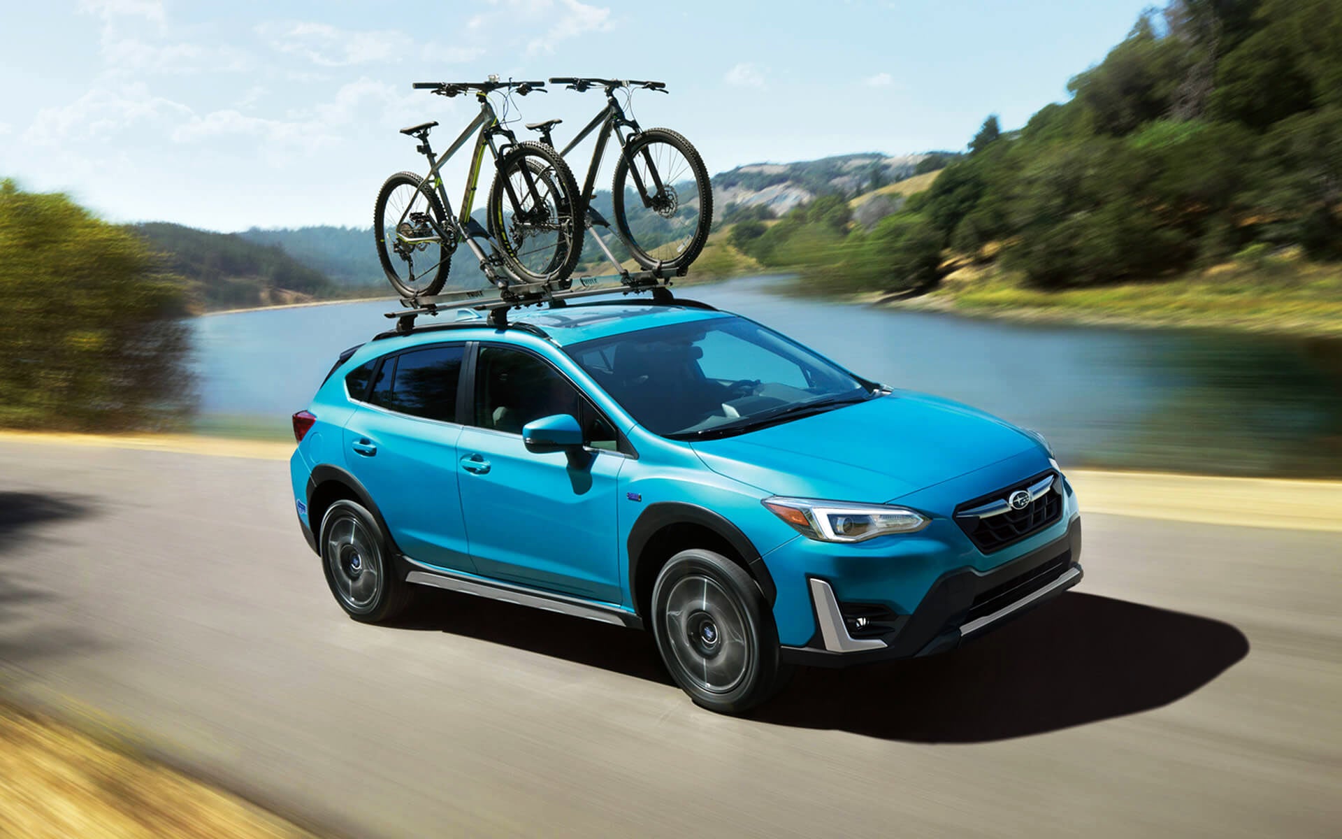 A blue Crosstrek Hybrid with two bicycles on its roof rack driving beside a river | Dean Team Subaru in Ballwin MO
